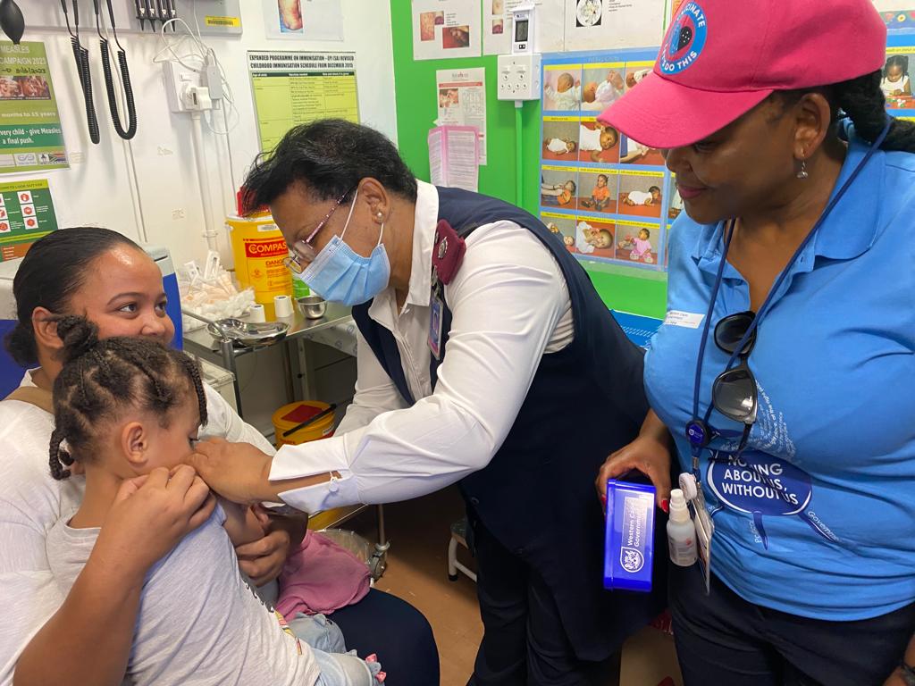 Three-year-old Ryleigh Du Preez receives her measles vaccine from nurse Gertrude Wessels at Kleinvlei Community Health Centre, while her mother Alyssa and Minister Mbombo look on.​​​​​​​